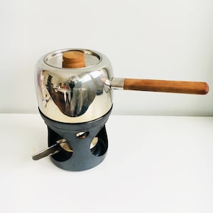 Puede incluir: Una olla de fondue de plata con un mango de madera y un soporte negro. La olla tiene una tapa con un pomo de madera.