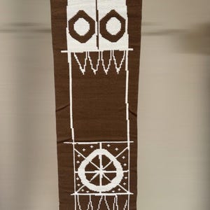 Puede incluir: Tapiz de pared tejido marrón y blanco con diseños geométricos. El textil rectangular presenta un patrón repetido de círculos, líneas y una forma de rueda. El borde inferior tiene flecos.