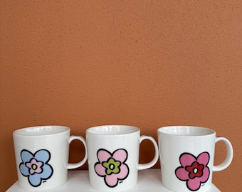 May include: Three white ceramic mugs with large, colorful flower designs. The flowers are outlined in black and feature shades of blue, pink, green, and red. Each mug has a curved handle and sits on a white surface against an orange background.