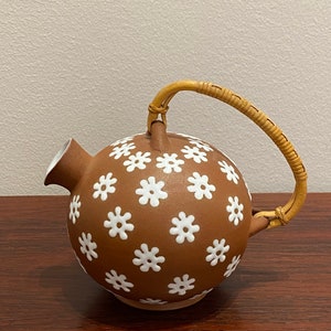 May include: A brown ceramic teapot with a white daisy pattern and a woven handle.
