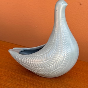 May include: A light blue ceramic bird planter with a textured, woven design. The planter is shaped like a bird with a long neck and a small head.