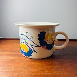 Könnte beinhalten: Cremefarbene Emaille-Tasse mit rundem Henkel. Die Tasse hat ein blaues Banner mit dem Wort "dom?" und zwei Blumendesigns in Blau, Gelb und Rosa. Die Tasse steht auf einer Holzoberfläche.