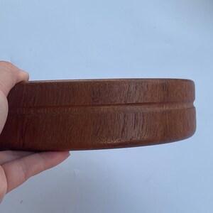 May include: A round, wooden bowl in a rich brown hue. The bowl features a smooth, polished surface and a simple, elegant design. The bowl is held by a hand against a white backdrop.