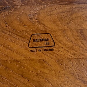 May include: Close-up of a wooden surface with a natural grain pattern. The wood is a warm brown colour. The text "BACKMAN -69- MADE IN FINLAND" is printed in black within a black outlined shape.