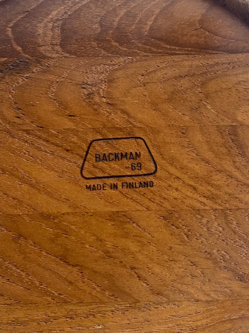 May include: Close-up of a wooden surface with a natural grain pattern. The wood is a warm brown colour. The text "BACKMAN -69- MADE IN FINLAND" is printed in black within a black outlined shape.