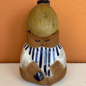 May include: A ceramic figurine of a person in a seated position. The figure has a light brown face and arms, with a yellow hair bun. The figure is wearing a white shirt with blue vertical stripes. The figurine is a decorative item.