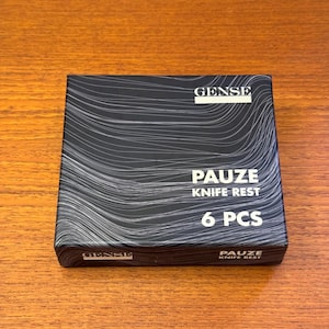May include: A black box with the text "GENSE" and "PAUZE KNIFE REST 6 PCS" in white. The box has a wavy, abstract design. The product is likely a set of knife rests.