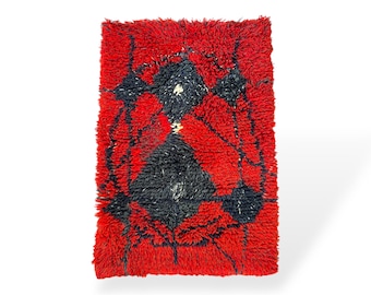 May include: A rectangular, shaggy rug with a vibrant red background. The rug features a central design in black, with white accents. The design appears to be abstract, with geometric shapes and lines. The rug is likely made of a soft, textured material.