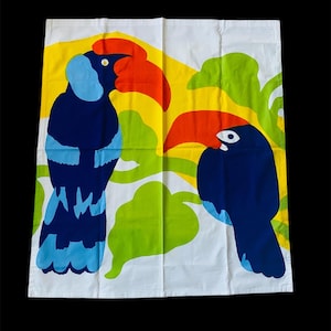 May include: A white fabric with a colorful design of two toucans with orange beaks and blue bodies. The toucans are surrounded by green and yellow leaves.