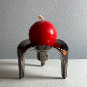 Puede incluir: Una vela roja se encuentra sobre un candelero de plata con tres patas. El candelero tiene un diseño único y moderno.