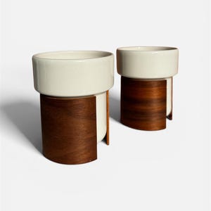 Op de afbeelding: Twee moderne keramische plantenbakken. Elke plantenbak heeft een witte, cilindrische bovenkant en een donkerbruine, gebogen houten basis. De plantenbakken hebben een minimalistisch ontwerp, geschikt voor kamerplanten of decoratief gebruik.