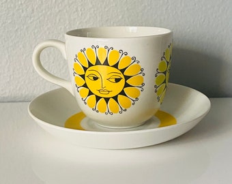 May include: A white ceramic teacup and saucer with a yellow sun design. The sun has a face with closed eyes and a smiling mouth.