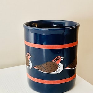 Peut inclure: Mug cylindrique en céramique bleu marine avec une bande corail et des illustrations d'oiseaux. Les oiseaux ont des détails marron, blancs et rouges. La tasse est de taille standard.
