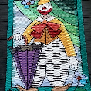 Peut inclure: Une tapisserie murale en point d'aiguille colorée représentant un clown tenant un parapluie. Le clown porte une tenue rayée jaune et blanche avec un nœud papillon rouge. L'arrière-plan est un paysage vert et blanc avec des fleurs.