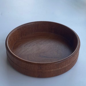 May include: A circular wooden bowl in a deep brown hue. The bowl features a straightforward design with a subtly elevated rim and a smooth inner surface. The wood grain is apparent, enhancing its natural appeal. Suitable for serving or display, this bowl is a versatile item.