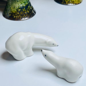 Puede incluir: Dos figuritas de oso polar de cerámica blanca. El oso más grande está de pie y mirando hacia la derecha. El oso más pequeño está sentado y mirando hacia arriba al oso más grande.