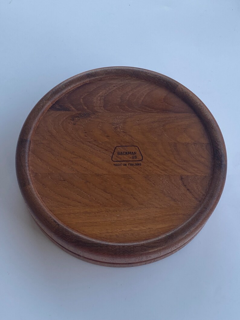 May include: A circular wooden tray with a raised rim, in a warm brown hue. The wood grain is clearly visible. The text "BACKMAN -89- MADE IN FINLAND" is stamped on the tray, indicating its origin.