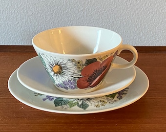 May include: A white teacup and saucer set with a floral pattern featuring red poppies, yellow daisies, and purple flowers. The set is on a wooden surface.