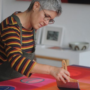 May include: A person paints a large abstract canvas with a wide brush. The canvas is red, orange, and blue with geometric shapes. The person is wearing a striped shirt and a green apron.