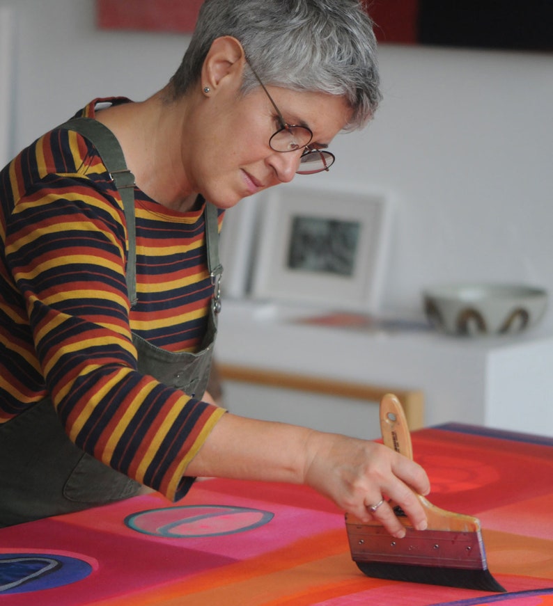 May include: A person paints a large abstract canvas with a wide brush. The canvas is red, orange, and blue with geometric shapes. The person is wearing a striped shirt and a green apron.