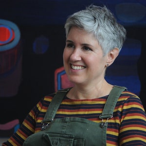May include: A woman with short, gray hair is smiling and looking to the right. She is wearing a striped long-sleeved shirt with a green overall.