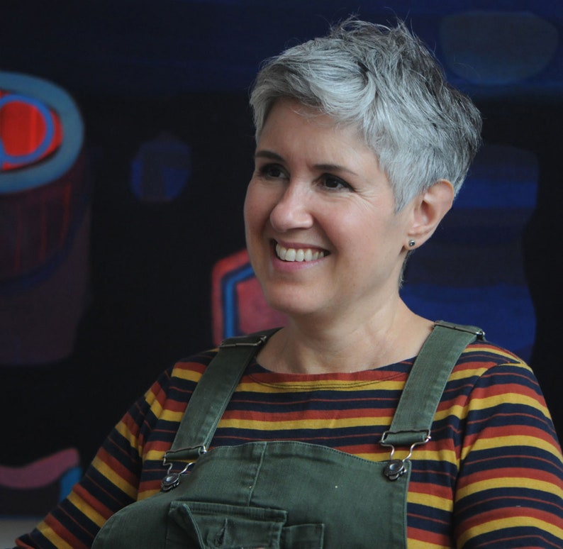 May include: A woman with short, gray hair is smiling and looking to the right. She is wearing a striped long-sleeved shirt with a green overall.
