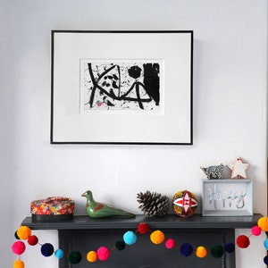 May include: A black and white abstract art print in a black frame hangs above a fireplace mantel. The mantel is decorated with a colorful pom-pom garland, a green ceramic bird, a pine cone, a red and white patterned ball, and a sign that reads "Happy".
