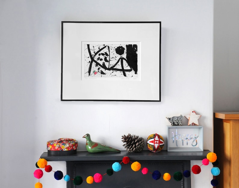 May include: A black and white abstract art print in a black frame hangs above a fireplace mantel. The mantel is decorated with a colorful pom-pom garland, a green ceramic bird, a pine cone, a red and white patterned ball, and a sign that reads "Happy".