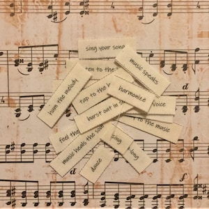 May include: A collection of small, rectangular tags with black text, scattered over a background of sheet music. The tags feature phrases like "sing your song," "hum the melody," and "music speaks." The background is a sepia-toned sheet music.