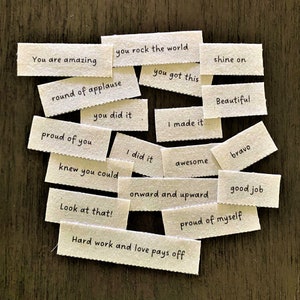 May include: A set of 16 small, white fabric tags with encouraging phrases written in black ink. The phrases include "You are amazing", "You rock the world", "Round of applause", "Proud of you", "You did it", "I did it", "Knew you could", "Look at that", "You got this", "I made it", "Awesome", "Onward and upward", "Shine on", "Beautiful", "Bravo", "Good job", "Proud of myself", and "Hard work and love pays off".