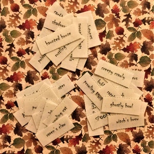 May include: A collection of small, white, rectangular fabric tags with black handwritten text. The tags are scattered on a brown background with a pattern of autumn leaves. The text on the tags includes words like "haunted house", "rustling", "creepy crawly", "ghostly howl", and "witch's brew".