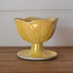 May include: A yellow ceramic planter shaped like a flower with a wide, shallow bowl and a pedestal base. The planter is sitting on a wooden surface.