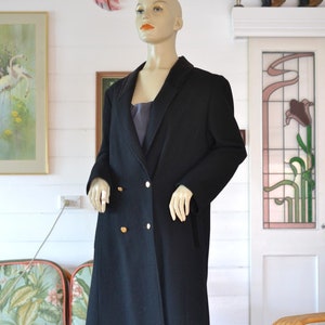 May include: A long black double-breasted coat with gold buttons. The coat is on a mannequin in a room with a floral patterned chair and a door with a stained glass window.