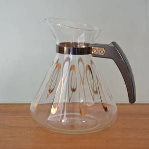 Può includere: Una caffettiera di vetro con manico nero e design geometrico dorato. La caffettiera ha una fascia argentata attorno alla parte superiore.