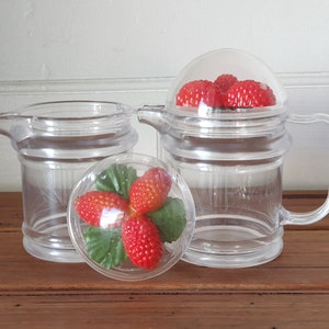 May include: Two clear plastic pitchers with lids. One pitcher has a lid with a red strawberry design. The other pitcher has a lid with a red strawberry design.