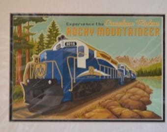 May include: A vintage travel poster featuring a blue and white train engine with the text "Experience the Canadian Rockies Rocky Mountaineer". The train is traveling through a mountainous landscape with trees and a river.