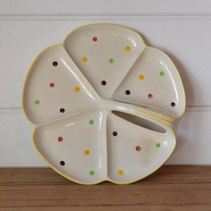 May include: A white ceramic serving dish shaped like a four-leaf clover with a yellow rim. The dish has four sections and is decorated with colorful polka dots in red, yellow, green, and purple.