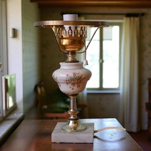 May include: A vintage table lamp with a floral design on a white ceramic base. The lamp features a brass-colored metal frame and a marble base. The lamp is on a wooden table.