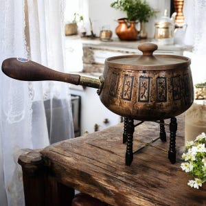 May include: An antique copper cooking pot with a wooden handle and lid, on a black stand. The pot has decorative rectangular panels with a dark design. The handle is dark brown with a brass fitting.