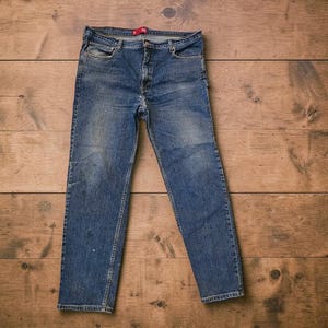 May include: A pair of blue denim jeans with a faded wash. The jeans have a classic five-pocket design, a button and zipper closure, and a red tag on the waistband. The jeans are laid flat on a wooden surface.