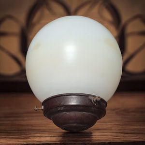 Puede incluir: Una lámpara de globo de vidrio blanco con una base de metal marrón. El globo es liso y tiene un ligero tono amarillo. La base tiene un diseño decorativo.