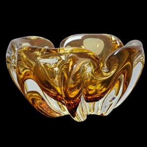 May include: An amber and clear glass bowl with a distinctive, flowing form. The bowl's design is reminiscent of a flower or natural form, with a glossy finish. The glass has a swirling, textured appearance. The bowl is a decorative item.