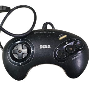 Pode incluir: Controlo Sega Mega Drive preto com um painel direcional, quatro botões de ação e um botão de arranque. O controlo tem a palavra "SEGA" impressa. O controlo está ligado a um cabo preto.