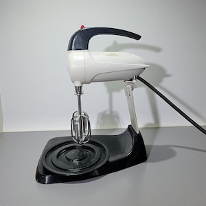 May include: A vintage hand mixer in cream and black, with a red button on top. The mixer has a chrome beater attachment and sits on a black base. The power cord is black. The mixer is designed for kitchen use.