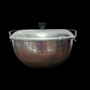May include: A large, round, metal cooking pot with a lid. The pot is silver-coloured with a black handle on the lid and two handles on the sides. The pot is set against a black background.