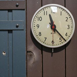 May include: A circular clock with a white frame and a cream face, featuring black hands and a red second hand. It's mounted on a brown wooden wall next to a blue wooden panel. The time displayed is roughly 11:20.