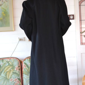 May include: A long black coat with a collar and long sleeves. The coat is made of a smooth fabric and has a simple design.