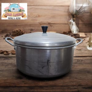 Peut inclure: Une casserole en aluminium argentée avec un couvercle et deux poignées. La casserole a un bouton noir sur le couvercle.