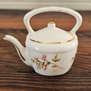 May include: A miniature white ceramic teapot featuring a delicate floral pattern with pink blossoms and green leaves. The teapot has a gold-trimmed lid and spout, a curved handle, and a rounded body, presenting a classic design for a collection.