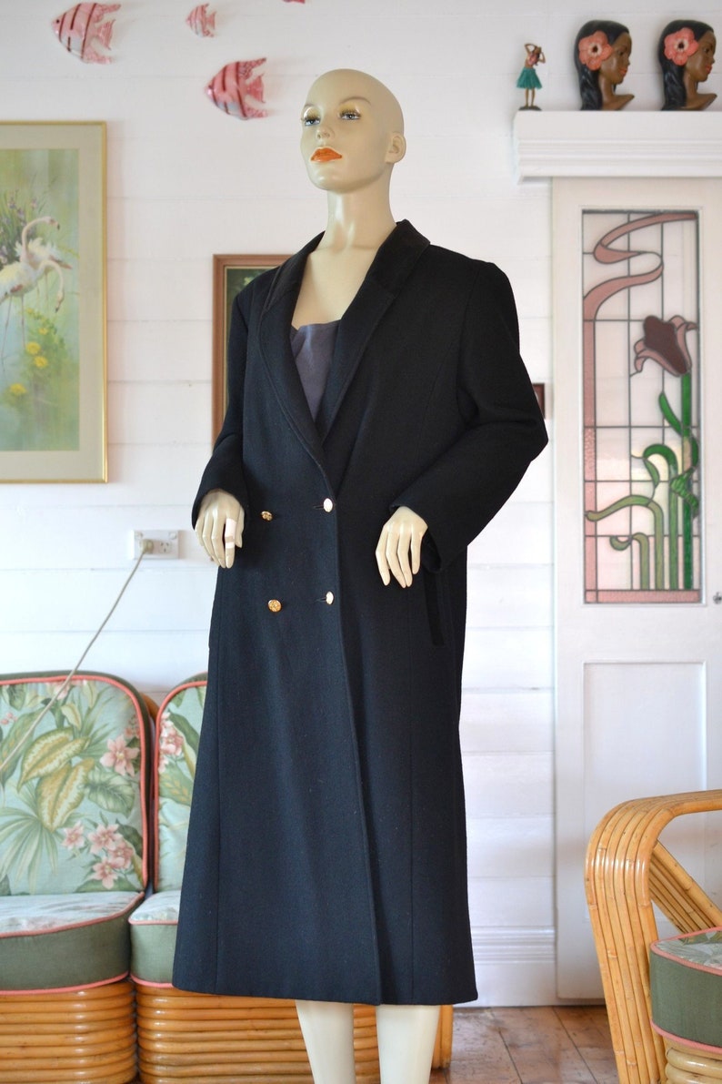 May include: A long black double-breasted coat with gold buttons. The coat is on a mannequin in a room with a floral patterned chair and a door with a stained glass window.